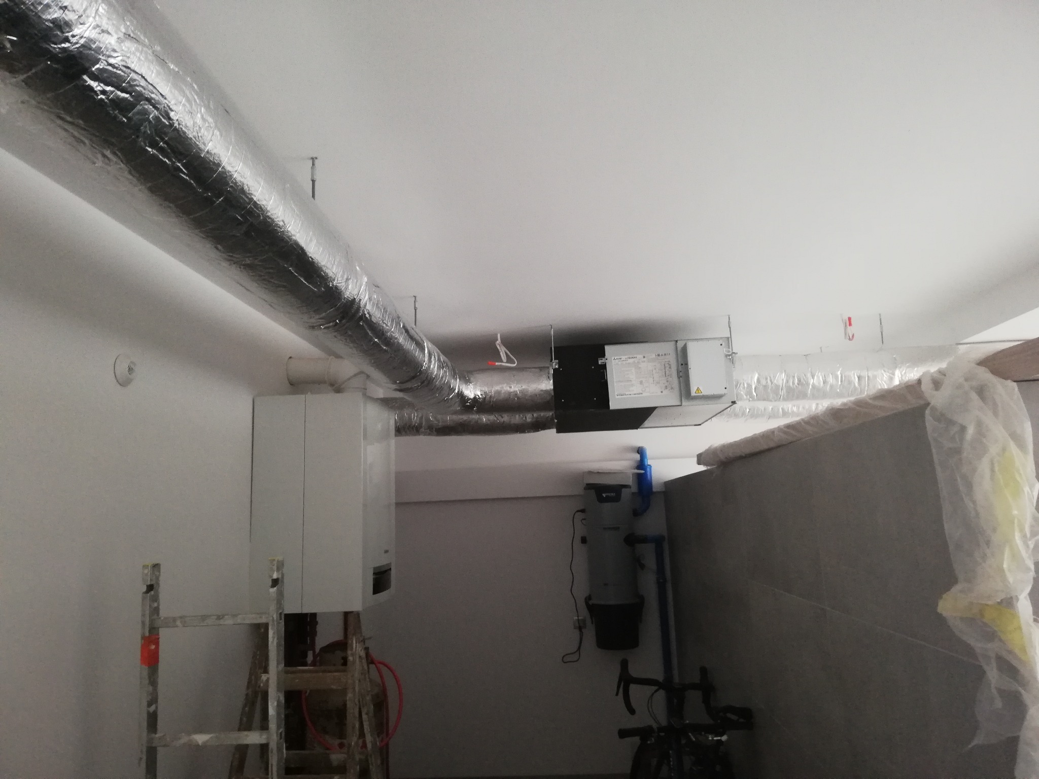 Szare, izolowane kanały wentylacyjne pod sufitem z zamontowaną centralą wentylacyjną, widoczny drabina i bojler w pomieszczeniu w trakcie prac wykończeniowych.