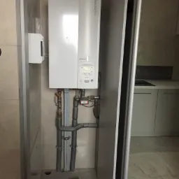 Biały piec grzewczy Atlantic zamontowany w zabudowie, widoczne szare rury instalacyjne i fragment kuchni w tle.