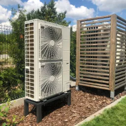Biała pompa ciepła typu split na czarnym metalowym stelażu, obok drewniana osłona na inną jednostkę zewnętrzną, na tle zieleni i błękitnego nieba z chmurami.