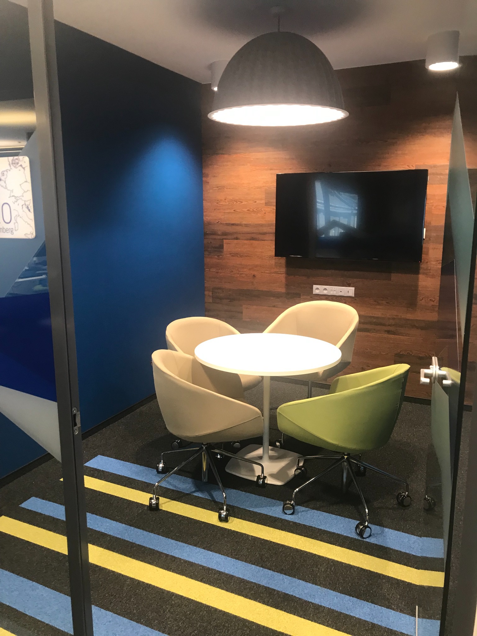 Kącik biurowy z okrągłym stołem, czterema krzesłami w odcieniach beżu i zieleni, dywanem w pasy, drewnianą ścianą z telewizorem i lampą sufitową