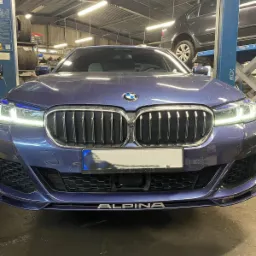 Przód niebieskiego BMW Alpina w warsztacie samochodowym, widoczny podnośnik z innym autem w tle oraz opony na półce.