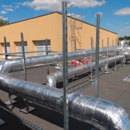 Izolowane rury wentylacyjne na dachu budynku komercyjnego, wsparte na metalowej konstrukcji, z widoczną drabiną dostępową i fragmentem instalacji. Widoczne niebo z chmurami i fragment budynku w tle.