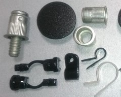 Zestaw różnorodnych metalowych i plastikowych elementów złącznych, w tym śruba z radełkowaną główką, tulejki, czarne i białe uchwyty kablowe oraz czarna zaślepka, ułożonych na jasnym tle.