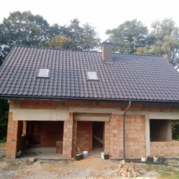 Niedokończony budynek z cegły z zamontowanym, ciemnoszarym dachem z dwoma oknami dachowymi i kominem, widoczne materiały budowlane na zewnątrz.