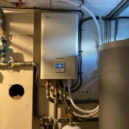 Biała pompa ciepła LG Therma V zamontowana na ścianie w piwnicy, widoczne podłączenia hydrauliczne i zbiornik buforowy, oświetlenie górne