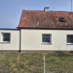 SES Group Fotowoltaika, Pompy Ciepła, Kotły, Klimatyzacja, Rekuperacja, Termomodernizacja - Jasny budynek z czerwoną dachówką i trzema oknami, otoczony zielonym trawnikiem. Na trawniku widoczny mały, czarny pies. Ujęcie w słoneczny dzień.