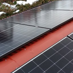 SES Group Fotowoltaika, Pompy Ciepła, Kotły, Klimatyzacja, Rekuperacja, Termomodernizacja - Panele fotowoltaiczne na czerwonym dachu domu, widok z góry, zieleń w tle. Montaż paneli słonecznych na dachu pokrytym blachą trapezową.
