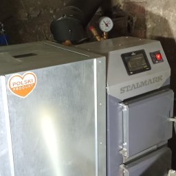SES Group Fotowoltaika, Pompy Ciepła, Kotły, Klimatyzacja, Rekuperacja, Termomodernizacja - Kocioł na paliwo stałe marki Stalmark z naklejką 'Polski Produkt' i czarnym kominem, zainstalowany w pomieszczeniu z ceglaną ścianą.