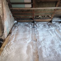 SES Group Fotowoltaika, Pompy Ciepła, Kotły, Klimatyzacja, Rekuperacja, Termomodernizacja - Izolacja poddasza pianą PUR, widoczne drewniane elementy konstrukcyjne i fragment ściany. Piana pokrywa całą powierzchnię, tworząc jednolitą warstwę izolacyjną.