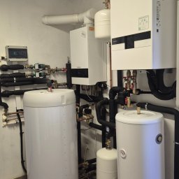 SES Group Fotowoltaika, Pompy Ciepła, Kotły, Klimatyzacja, Rekuperacja, Termomodernizacja - Nowoczesna kotłownia z dwoma białymi zasobnikami ciepłej wody i dwoma jednostkami grzewczymi, połączonymi rozbudowaną instalacją rur z izolacją i licznymi zaworami, na tle jasnych ścian.