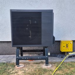 SES Group Fotowoltaika, Pompy Ciepła, Kotły, Klimatyzacja, Rekuperacja, Termomodernizacja - Zainstalowana pompa ciepła Panasonic Aquarea na metalowym stelażu, stojąca przy jasnej elewacji budynku, obok żółtej skrzynki gazowej.