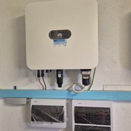 SES Group Fotowoltaika, Pompy Ciepła, Kotły, Klimatyzacja, Rekuperacja, Termomodernizacja - Biały inwerter Huawei z naklejką Fundusze Europejskie, zamontowany na jasnej ścianie. Widoczne podłączenia i dwie nowe rozdzielnice elektryczne w folii.