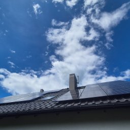 SES Group Fotowoltaika, Pompy Ciepła, Kotły, Klimatyzacja, Rekuperacja, Termomodernizacja - Błyszczące panele słoneczne na ciemnym, spadzistym dachu z dachówki, widoczne od dołu na tle błękitnego nieba z białymi chmurami. Obok komin i okno dachowe.