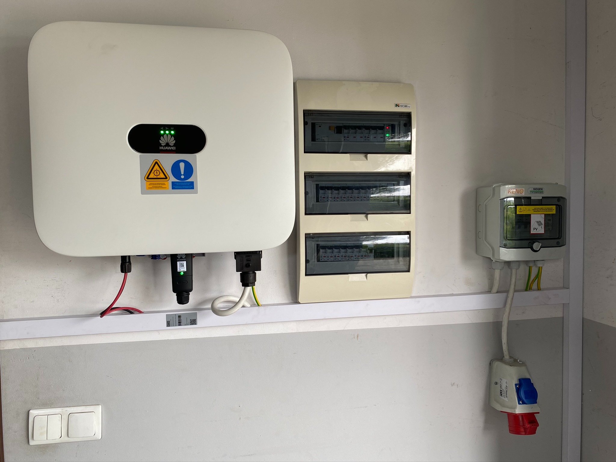 Biały inwerter Huawei z zielonymi diodami LED, obok skrzynka z bezpiecznikami i gniazdo siłowe na szarej ścianie.