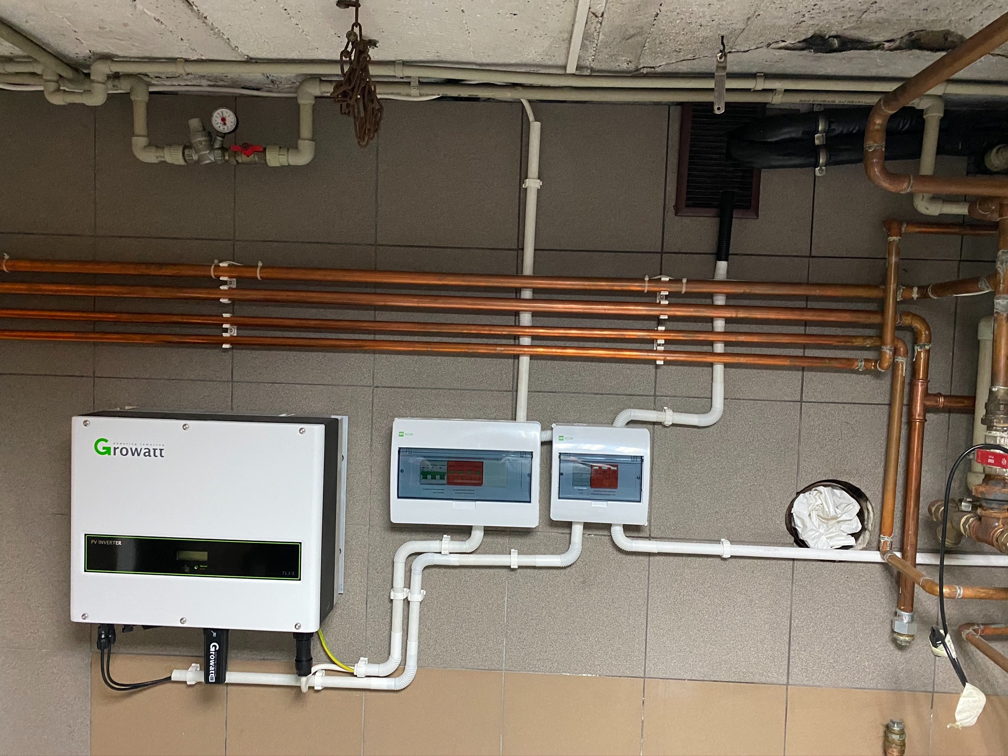 Inwerter fotowoltaiczny Growatt i rozdzielnice elektryczne na tle instalacji hydraulicznej z miedzianymi rurami, widoczne przewody i rury prowadzone po ścianie z szarych płytek.