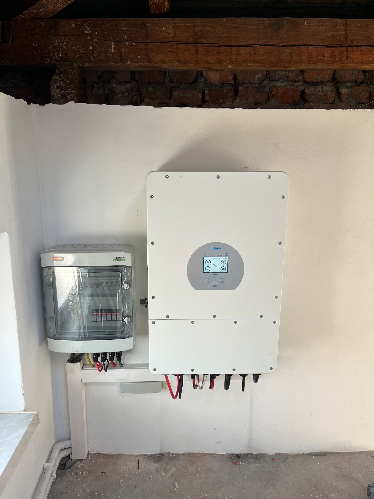 Biały falownik i skrzynka rozdzielcza zamontowane na ścianie wewnątrz budynku, widoczne kable i przewody, surowy stan wykończenia wnętrza, drewniana konstrukcja stropu.