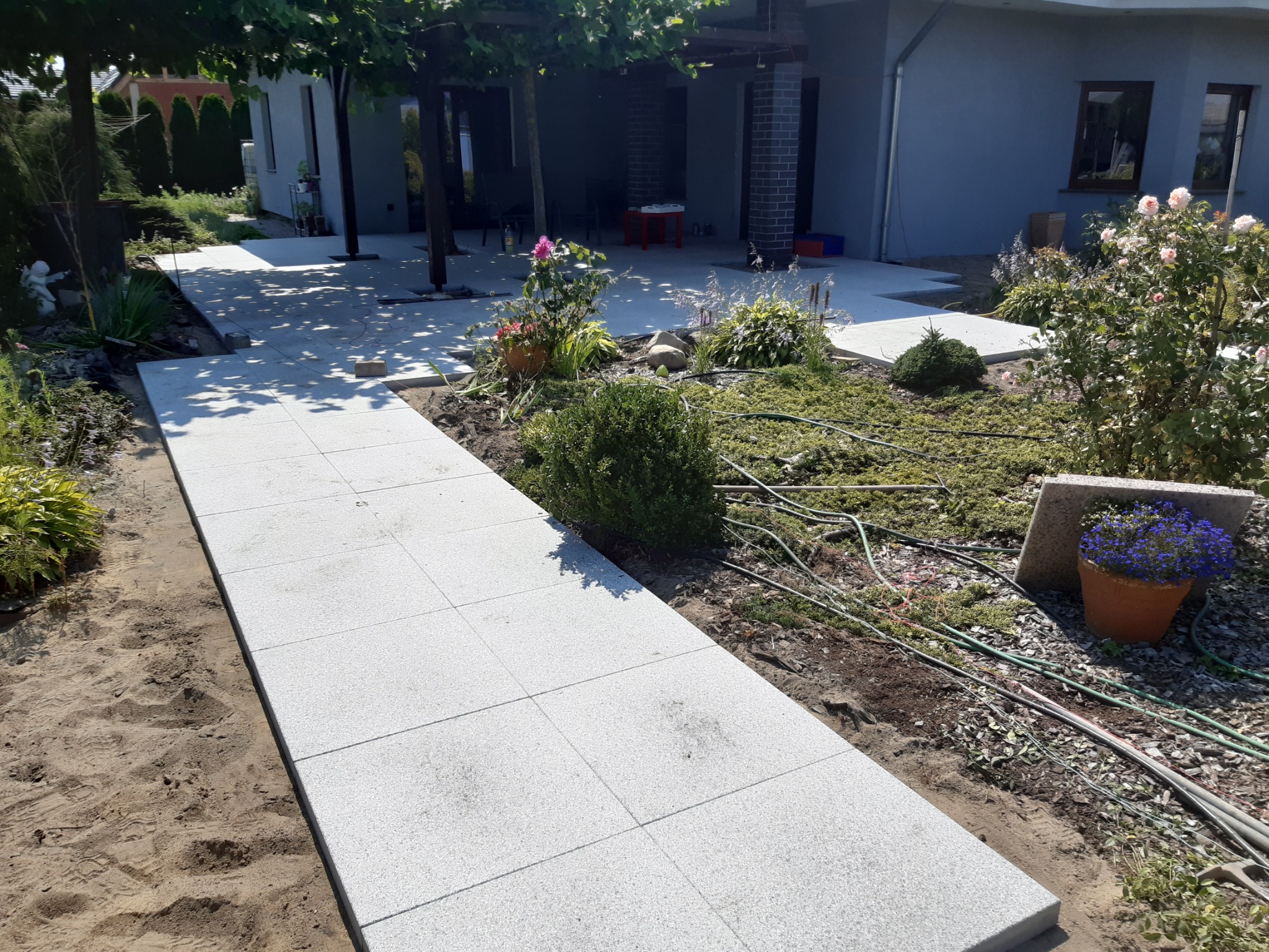 Ścieżka z jasnych, kwadratowych płyt prowadząca przez ogród w kierunku domu z zadaszonym tarasem, widoczne rośliny ozdobne i elementy instalacji nawadniającej.