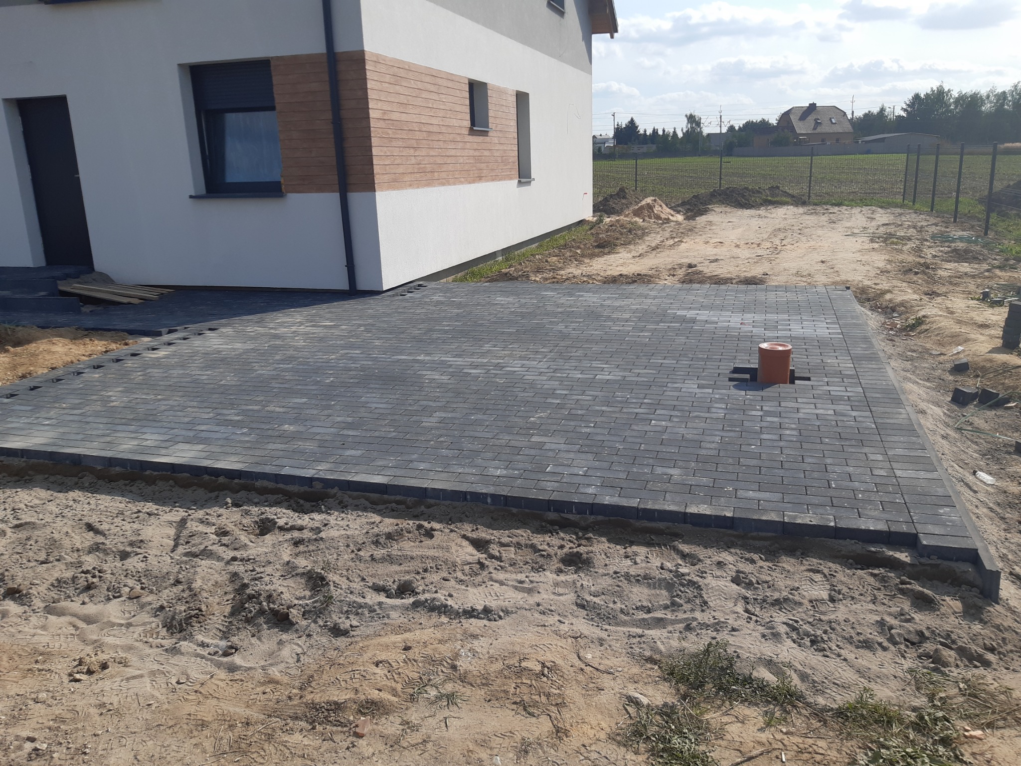 Układanie szarej kostki brukowej w prostokątnym wzorze obok nowo wybudowanego domu z elementami drewnianej elewacji, widoczne ślady pracy w postaci piasku i rury wentylacyjnej.