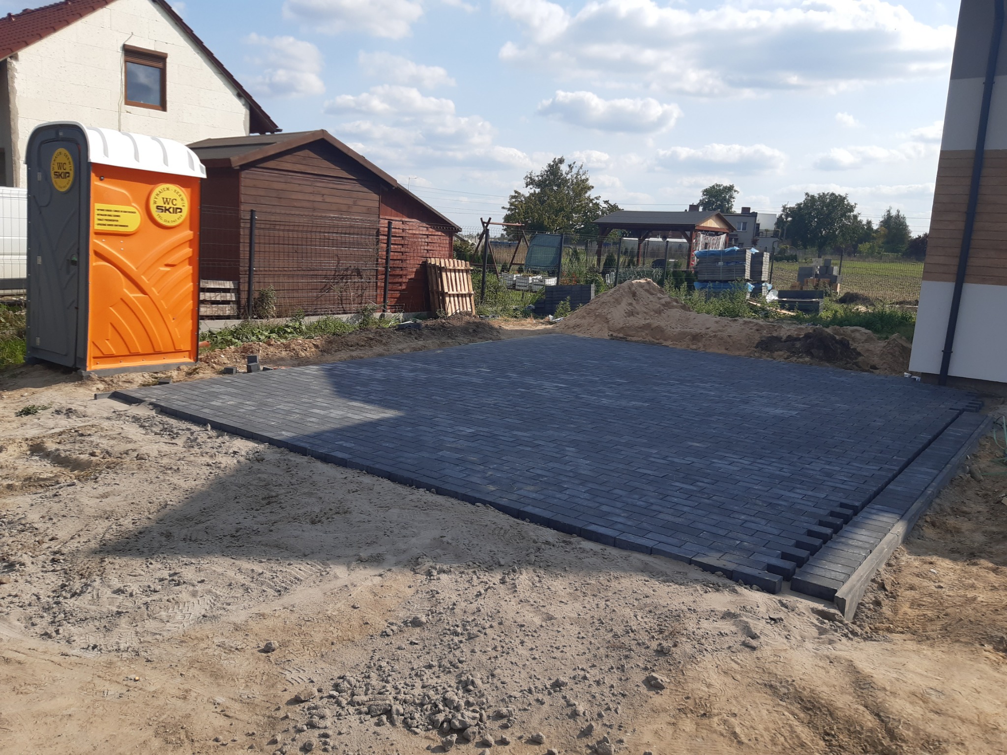 Częściowo ułożona, ciemnoszara kostka brukowa na piaszczystym podłożu, z widoczną przenośną toaletą WC Skip w tle.