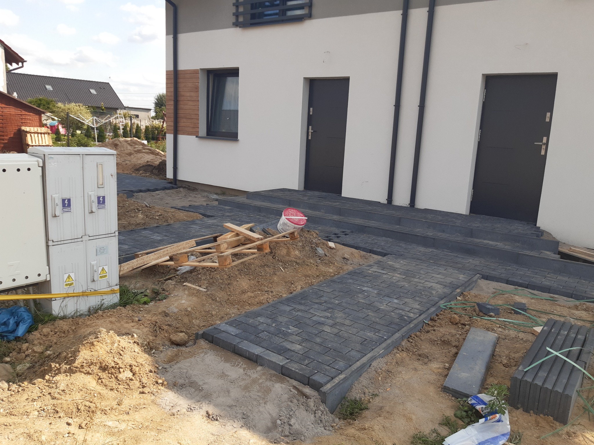 Częściowo ułożona, szara kostka brukowa prowadząca do dwóch wejść do bliźniaka; widoczne palety, piasek i skrzynka elektryczna.