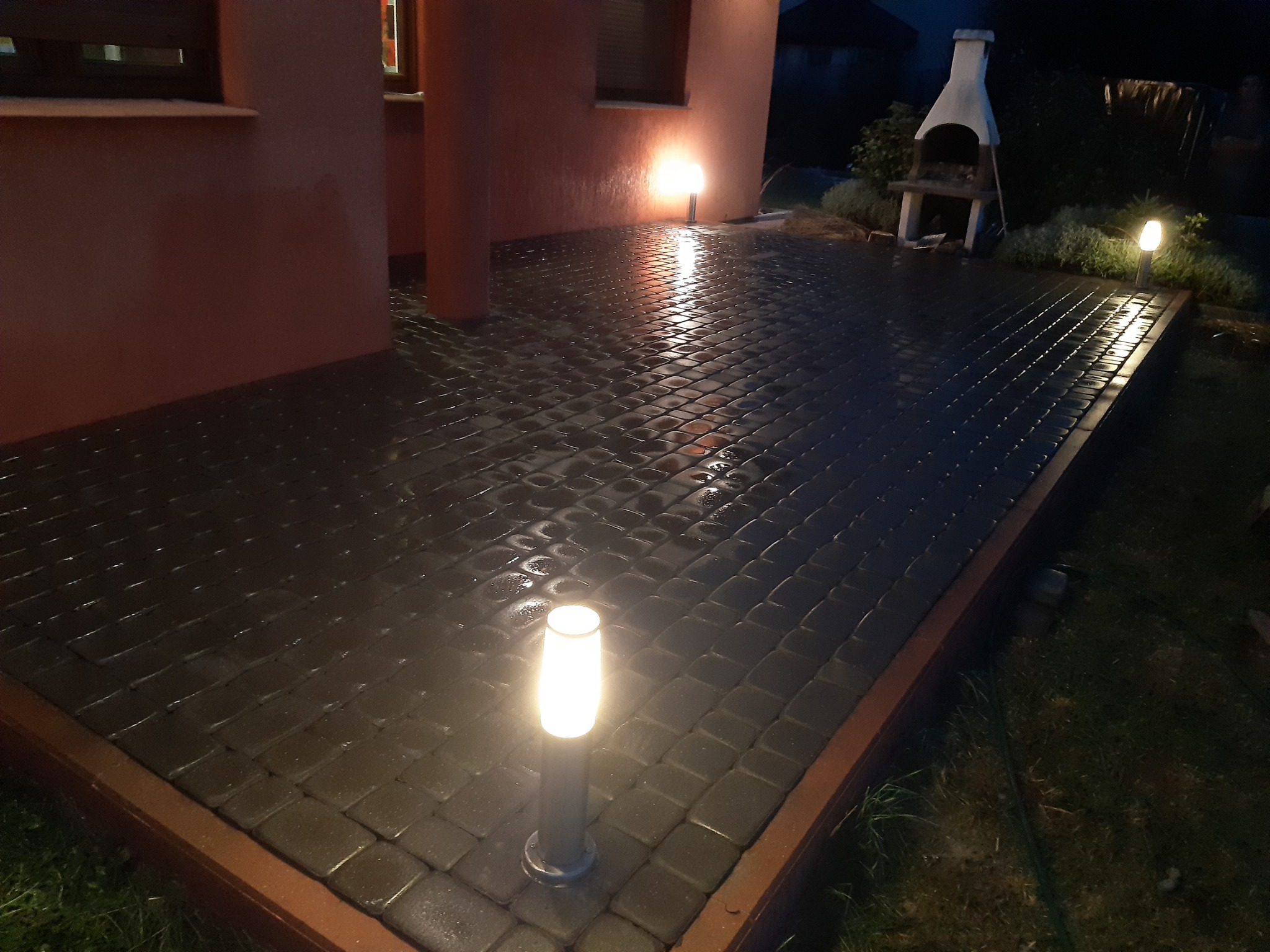 Ułożona szara kostka brukowa na posesji, oświetlona lampami ogrodowymi, widoczny budynek i grill w tle, wieczorne oświetlenie.