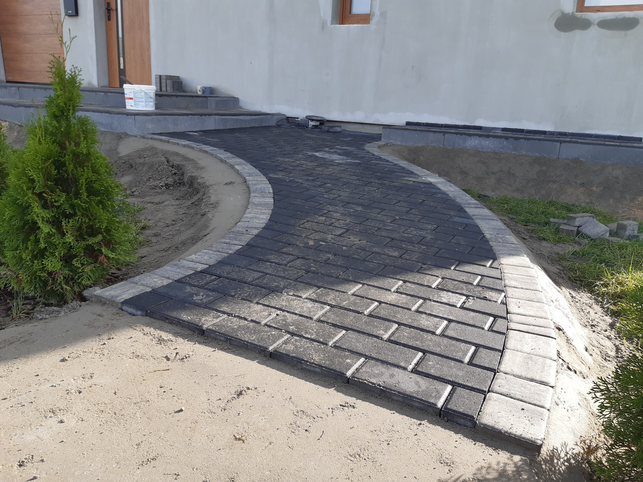 Zakrzywiona ścieżka z ciemnej kostki brukowej obrzeżona jasnoszarą kostką, prowadząca do drzwi wejściowych domu z białymi ścianami, widoczne tuje i piasek.