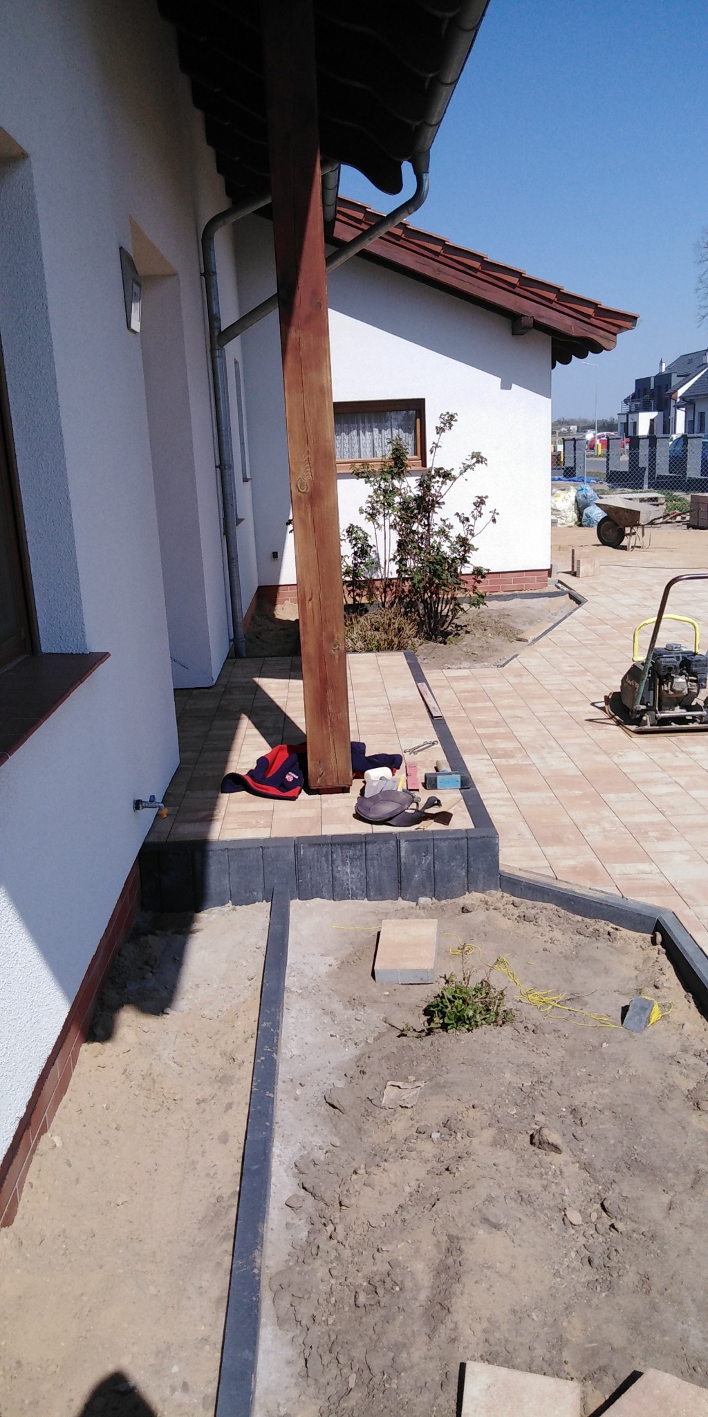 Układanie kostki brukowej: Widok na częściowo ułożoną kostkę brukową w odcieniach beżu, z widocznymi obrzeżami i przygotowanym podłożem. Na kostce leżą narzędzia, ubrania robocze i zagęszczarka.