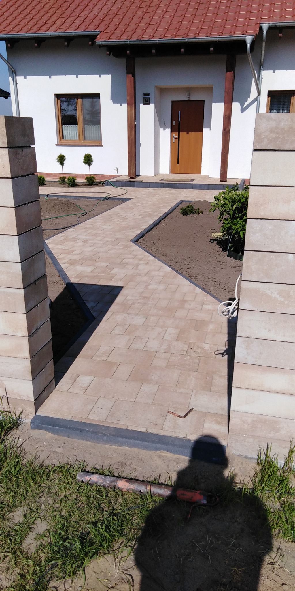 Ułożona ścieżka z kostki brukowej prowadząca do domu z czerwonym dachem, widoczne słupki ogrodzeniowe z niezakończonymi spoinami i cień osoby fotografującej.
