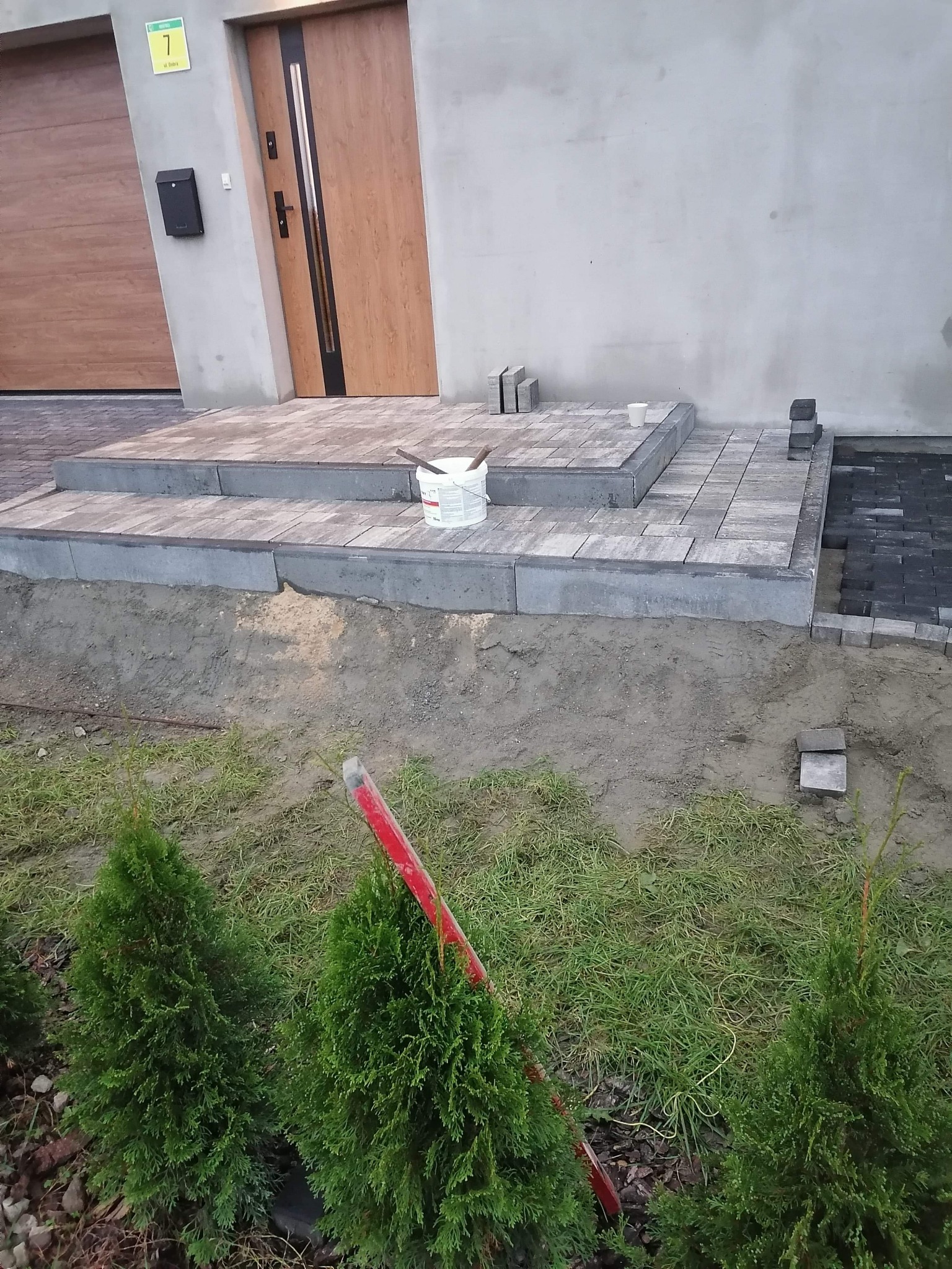 Układanie kostki brukowej przed wejściem do domu: widoczne dwustopniowe schody z szarej kostki, białe wiaderko po farbie, częściowo ułożona nawierzchnia z ciemnej kostki, fragment elewacji...
