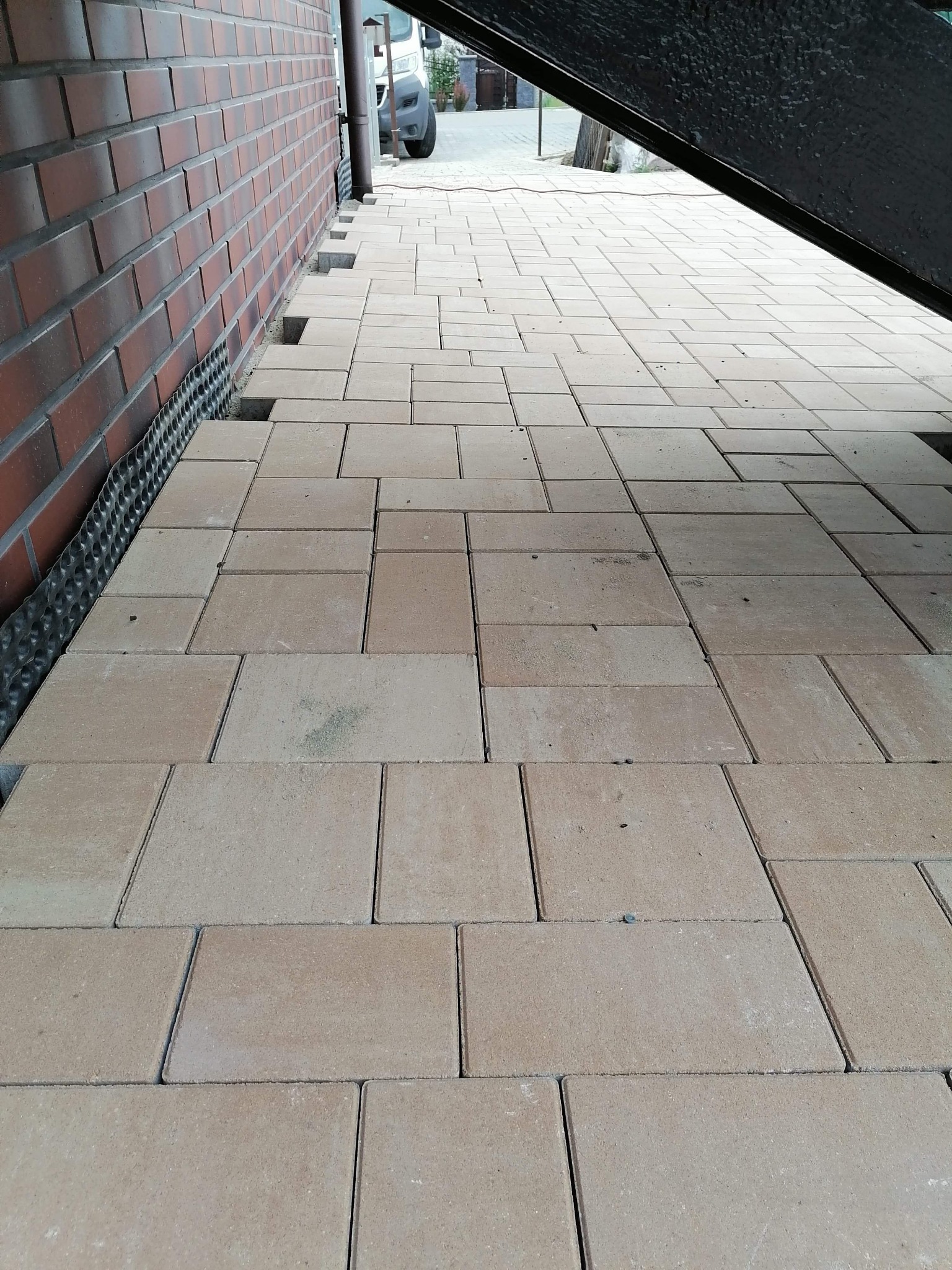 Fragment podjazdu z beżowej kostki, częściowo ułożonej, z widoczną membraną kubełkową przy fundamencie budynku z czerwonej cegły.