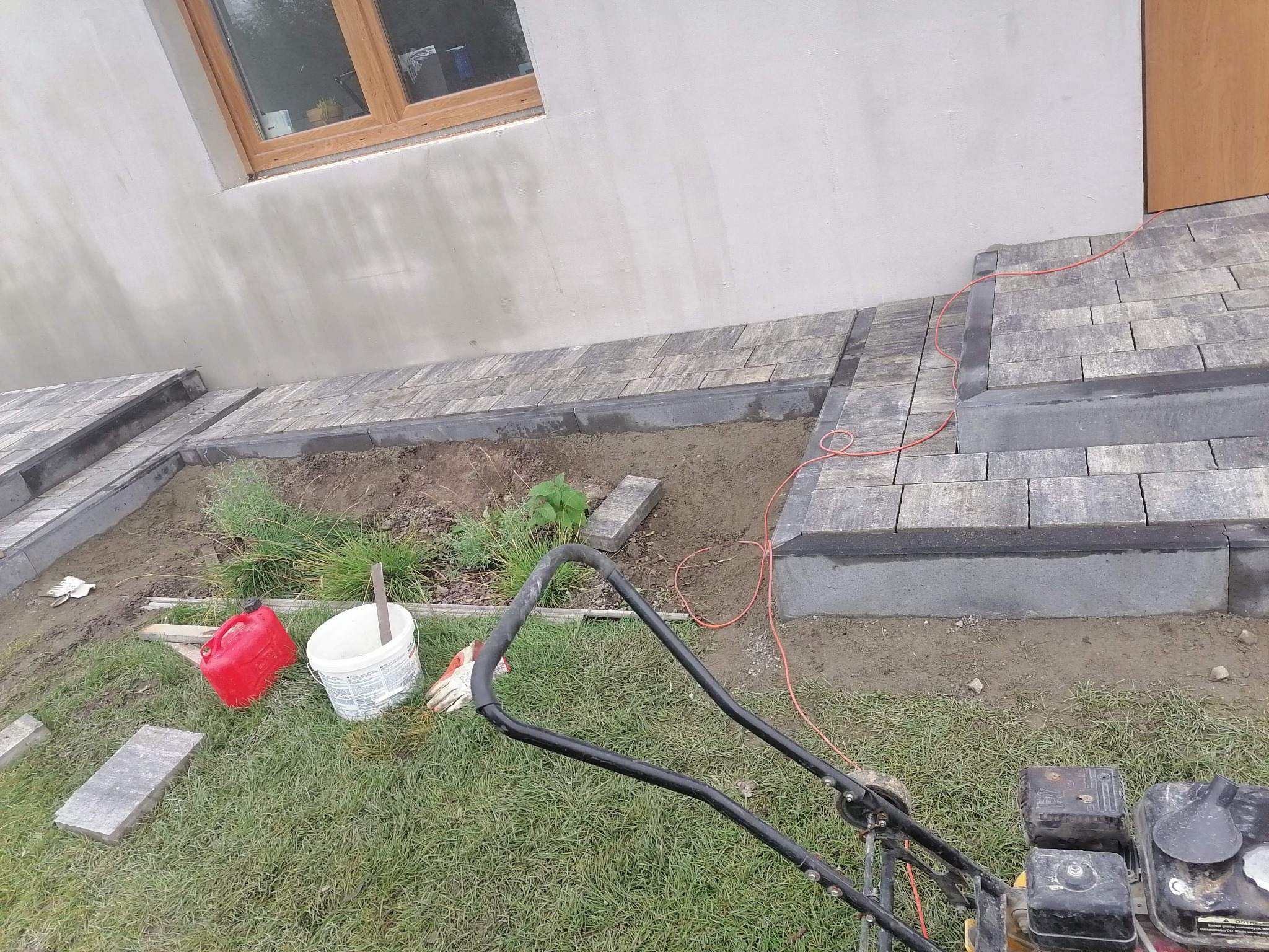 Świeżo ułożona ścieżka z kostki brukowej prowadząca do drzwi, z widoczną ubijarką mechaniczną, kanistrem i wiadrem na trawniku, obok budynku z oknem.