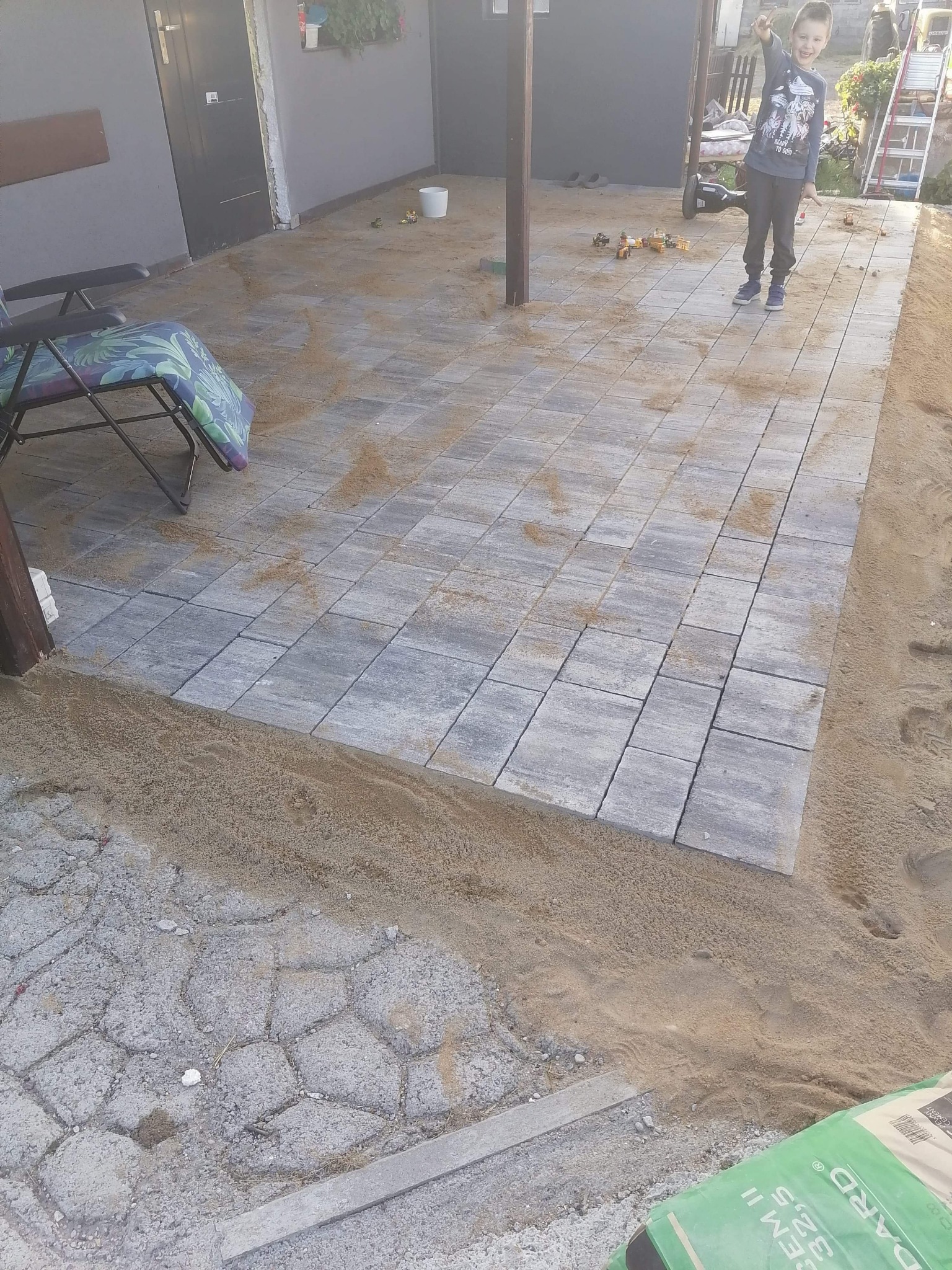 Niemal ukończony taras z szarych kostek brukowych, zasypany piaskiem, zabawki budowlane i uśmiechnięty chłopiec w tle, widoczny fragment worka z cementem.