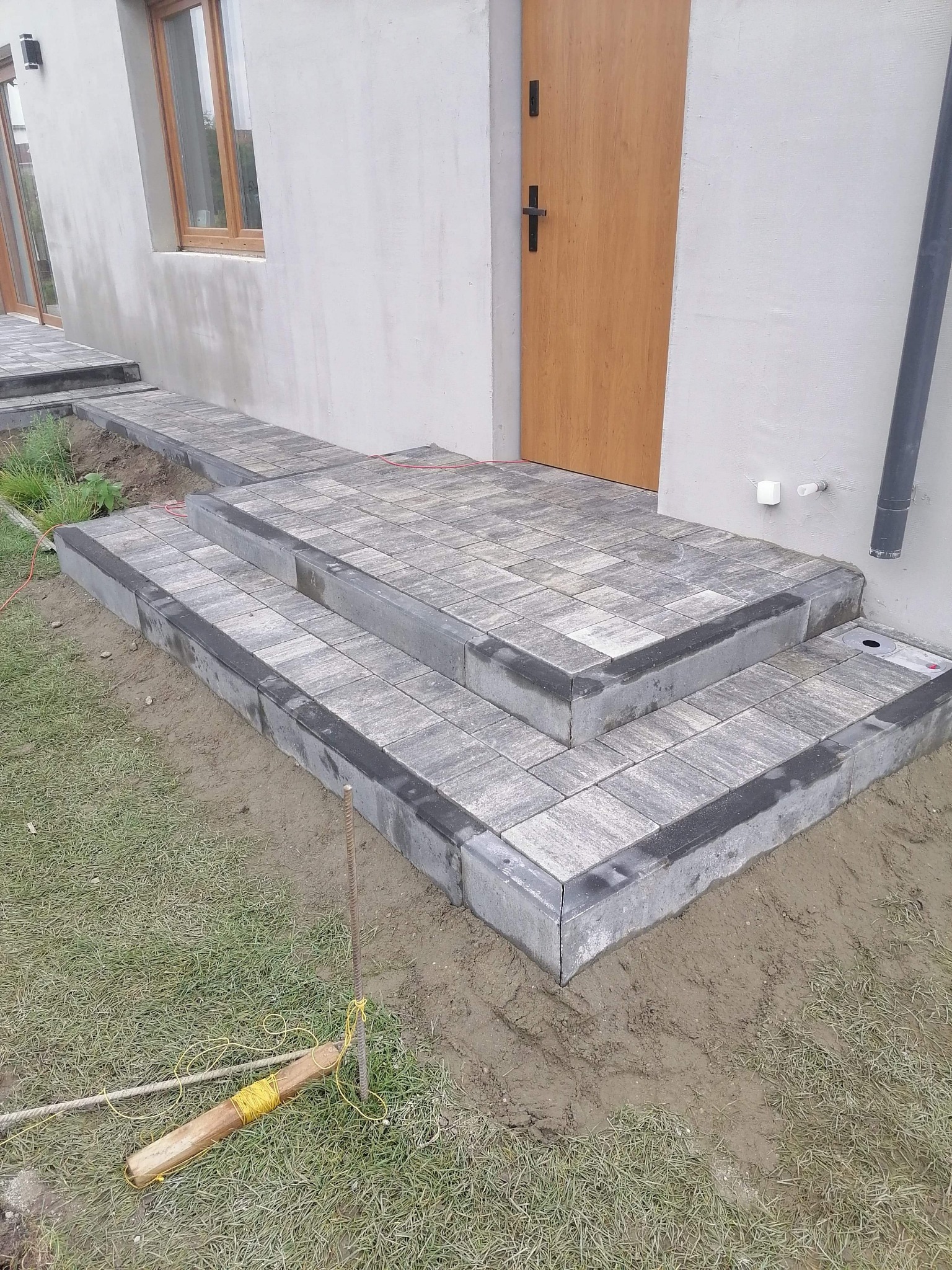 Schody wejściowe wyłożone szarą kostką brukową prowadzące do drewnianych drzwi wejściowych, widoczne narzędzia brukarskie i fragment chodnika z kostki.