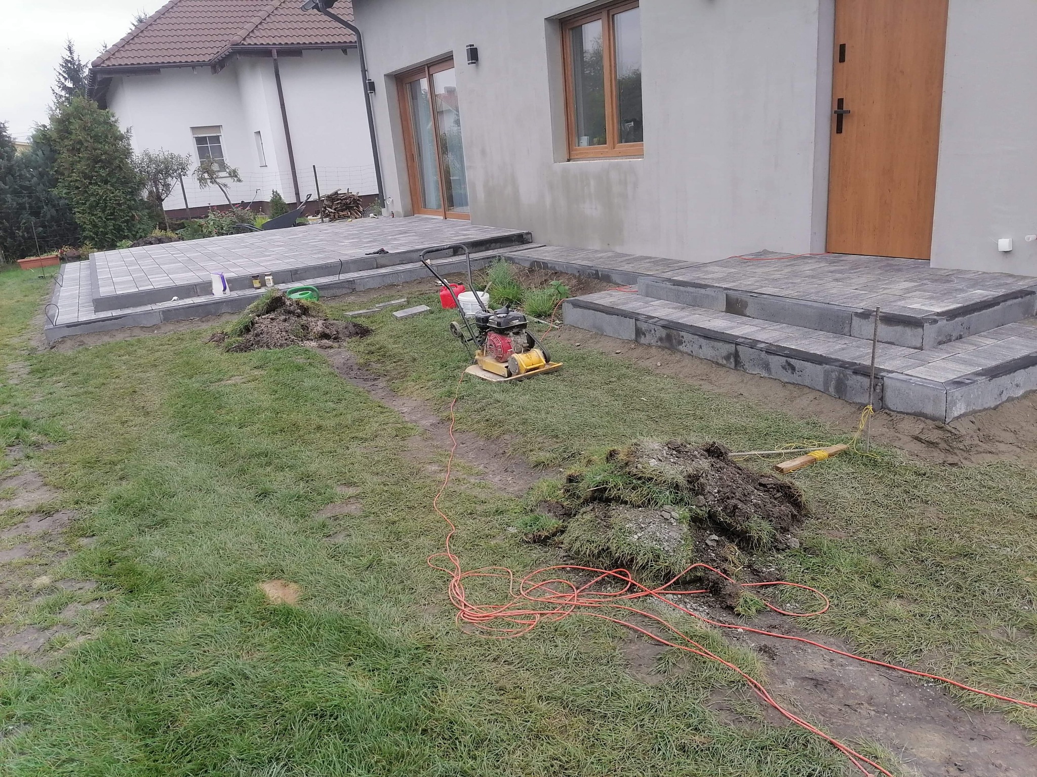 Etap prac: Ułożona kostka brukowa tworząca schody i taras przed domem, widoczna zagęszczarka i kable na trawniku.