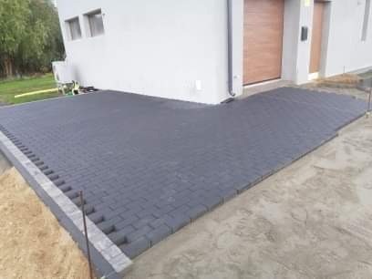 Ułożona, szara kostka brukowa w kształcie litery L przed budynkiem z brązową bramą garażową, widoczne obrzeża i piasek w spoinach.