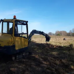 Żółta minikoparka Volvo na gąsienicach w trakcie pracy na polu, z widocznym świeżym wykopem i niebieskim niebem w tle.