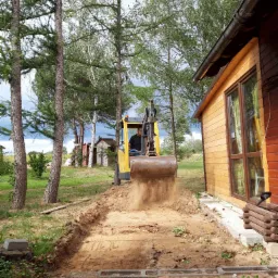 Żółta mini koparka w trakcie wykopu wąskiego rowu pod fundamenty w Zielonej Górze, z ziemią unoszącą się z łyżki, widok z perspektywy niskiej ceglanej ścieżki.