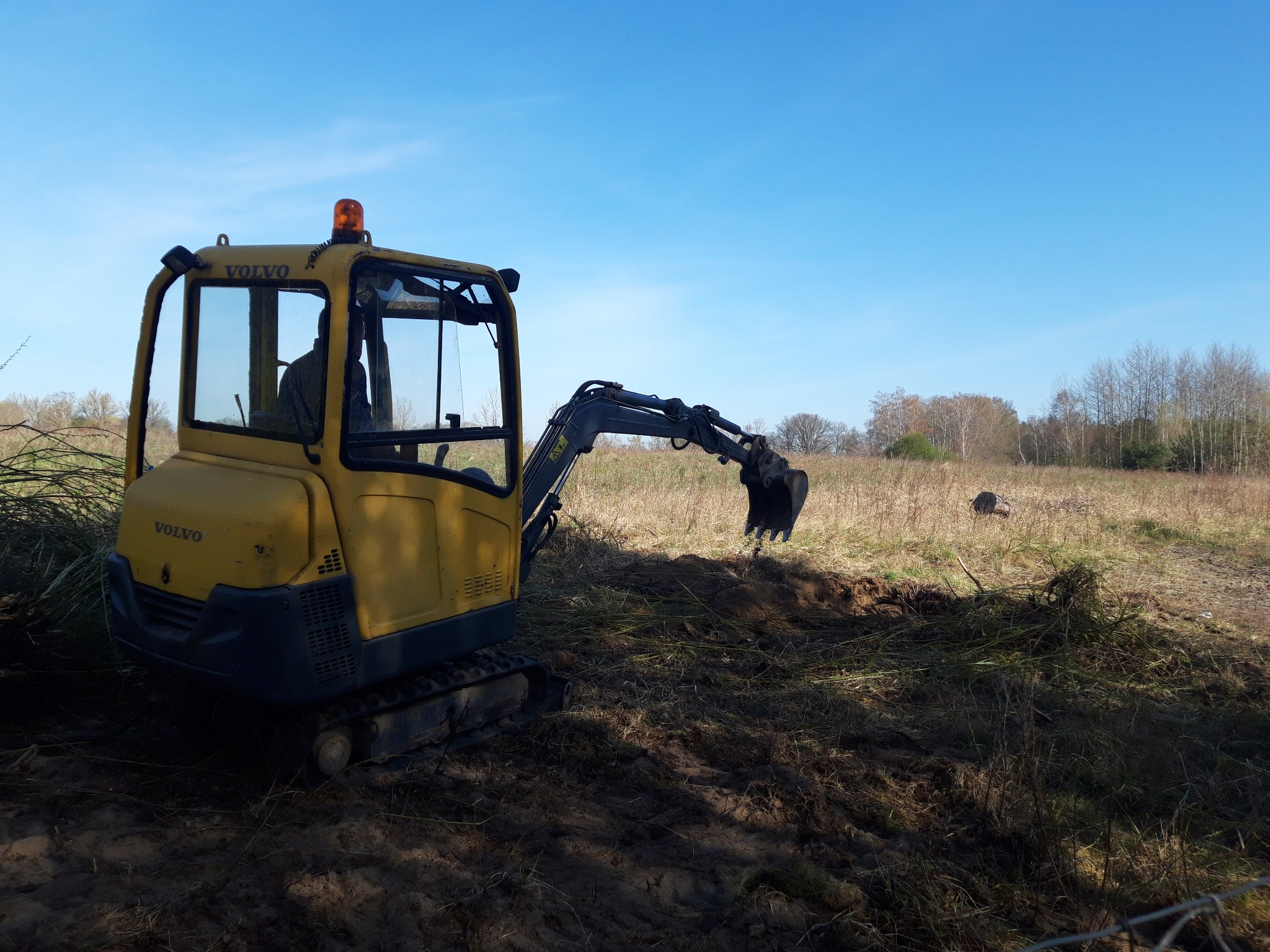 Żółta minikoparka Volvo na gąsienicach w trakcie pracy na polu, z widocznym świeżym wykopem i niebieskim niebem w tle.