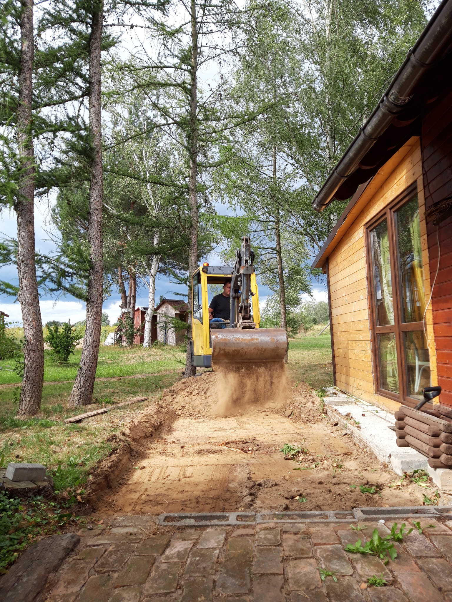 Żółta mini koparka w trakcie wykopu wąskiego rowu pod fundamenty w Zielonej Górze, z ziemią unoszącą się z łyżki, widok z perspektywy niskiej ceglanej ścieżki.