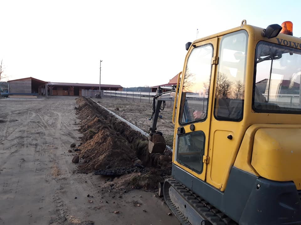 Żółta minikoparka Volvo podczas wykopu wąskiego rowu w ziemi, prawdopodobnie pod instalację, z budynkiem w tle, wczesnym rankiem.
