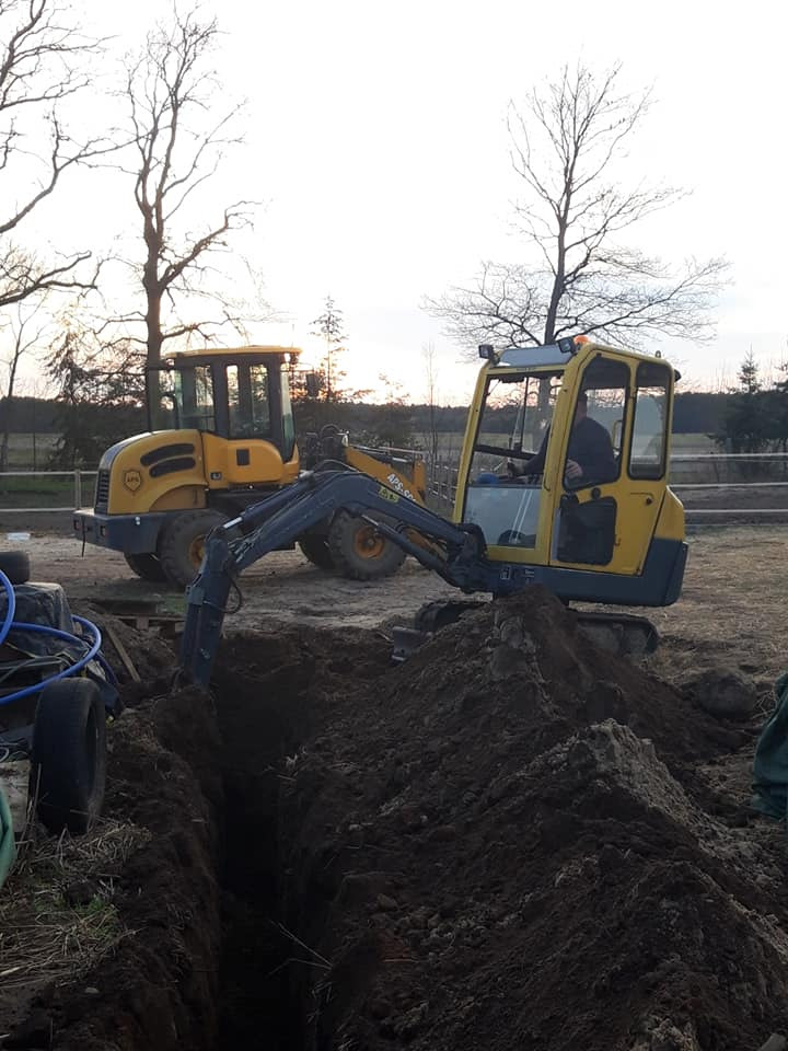 Żółty minikoparka i walec w trakcie prac ziemnych, wykop wąskiego rowu w ziemi, widoczne drzewa bez liści w tle.