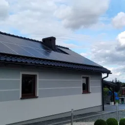 Dom jednorodzinny z panelami słonecznymi na dachu pokrytym czarną dach&oacute;wką, widok z ogrodu w słoneczny dzień.