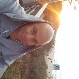 Autoportret osoby w szarej bluzie z kapturem i słuchawkami na tle plaży i zachodzącego słońca, widoczny znak z napisem 'Wejście' i literą 'G'.