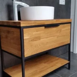 Łazienkowa szafka pod umywalkę z blatem i półką z jasnego drewna, czarne metalowe nogi, okrągła biała umywalka nablatowa, chromowana bateria, szare płytki na ścianie i podłodze.