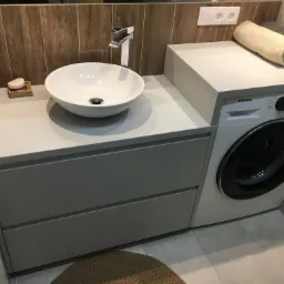 Nowoczesna łazienka z umywalką nablatową, szarą szafką z szufladami i pralką Samsung, uzupełniona o drewniane akcenty na ścianie i okrągły dywanik.
