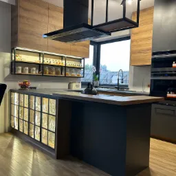 Nowoczesna kuchnia z wyspą, drewnianymi szafkami g&oacute;rnymi i przeszklonymi szafkami dolnymi z podświetleniem, czarny okap i metalowa konstrukcja nad wyspą.