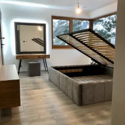 Nowoczesna sypialnia z podnoszonym łóżkiem tapicerowanym w odcieniach szarości, widoczny mechanizm podnoszenia stelaża, toaletka z podświetlanym lustrem i widok na zasypany śniegiem ogród za oknem.