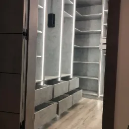 Wnętrze garderoby z szufladami i półkami, oświetlenie LED w pionowych panelach, wykończenie w odcieniach szarości, widok z perspektywy wejścia.