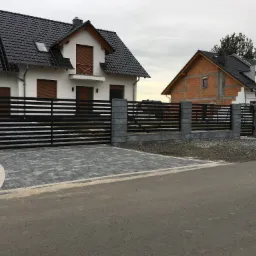 Nowoczesna, pozioma brama wjazdowa z czarnych paneli, osadzona między szarymi, betonowymi słupkami, prowadząca do posesji wyłożonej szarą kostką brukową, z widocznym w tle białym domem z ciemną...