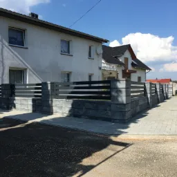 Nowoczesne ogrodzenie z paneli poziomych i betonowych słupków, widok na dom z białej cegły i niebieskie niebo z chmurami.